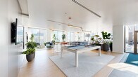 Located in Chicago's vibrant West Loop neighborhood at 123 N Desplaines St, this stylish amenity lounge offers a generously proportioned, open-plan layout flooded with natural light from expansive floor-to-ceiling windows that frame sweeping views of the city skyline. The light oak hardwood flooring and crisp white ceilings create a bright, airy atmosphere throughout the space.

The main area is anchored by a sleek pool table atop a soft area rug, surrounded by high-top bar tables and modern pedestal stools — ideal for social gatherings. Adjacent to this is a well-appointed bar counter with a bold navy blue island and white bar stools, positioned alongside full-height glass walls that provide direct access to a furnished outdoor terrace.

Multiple distinct lounge zones feature contemporary, modular seating in soft blue, green, and neutral tones, accented with colorful throw pillows and lush indoor greenery. A more intimate seating area with a round wood dining table and mint green chairs offers a relaxed setting for smaller groups.

Floor-to-ceiling glass partitions divide portions of the space while maintaining visual openness. Wall-mounted flat-screen televisions, pendant lighting with warm gold accents, and abundant tropical plants contribute to a polished yet welcoming ambiance throughout this exceptional urban retreat.