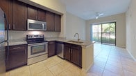 This single-floor residence offers a thoughtfully designed layout that seamlessly connects the kitchen, living area, and outdoor space. The kitchen is a standout feature, equipped with rich dark-espresso cabinetry extending to the ceiling, granite countertops, stainless steel appliances including an electric range, built-in microwave, dishwasher, and refrigerator. A peninsula with a double-basin sink and a brushed gold faucet serves as a natural divider between the kitchen and the open living area, enhancing the functional flow of the space. Light beige ceramic tile flooring runs throughout the kitchen and common areas, contributing to a clean, cohesive aesthetic. The living area is bright and airy, benefiting from a ceiling fan and a sliding glass door that opens to a private outdoor balcony with lush greenery views. The bedroom is generously sized, featuring soft wall-to-wall carpeting, a ceiling fan, and a window fitted with blinds that allows for natural light. A full bathroom completes the space, showcasing matching dark cabinetry with a granite-top vanity, a large mirror, and a tub and shower combination with tile surround. The neutral color palette and consistent finishes throughout create a cohesive and inviting atmosphere.