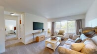 This single-floor residence offers a well-organized layout with a coastal-inspired aesthetic throughout. The living room serves as the heart of the space, featuring soft carpeting, a wall-mounted flat-screen television, and a large picture window that frames outdoor greenery while flooding the room with natural light. A sliding door leads to a balcony, further enhancing the connection to the outdoors. Neutral walls and warm, layered furnishings create a relaxed and inviting atmosphere.

Adjacent to the living area is a dedicated dining nook, defined by a pendant light overhead and accented with artwork, providing an intimate setting for meals. The galley-style kitchen features warm oak cabinetry, dark countertops, hardwood-style flooring, and a full suite of appliances including a range, refrigerator, and dishwasher. A dedicated laundry room with a full-size washer and dryer and overhead cabinetry adds everyday convenience.

The unit includes two carpeted bedrooms, each with closet space and natural light from well-placed windows. The bathroom offers a generous vanity with oak cabinetry and a wide mirror. Throughout, the space maintains a clean, comfortable character well-suited for everyday living.