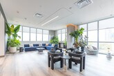 Located at 205 Queen St E in Brampton, Ontario, this contemporary amenity lounge spans a generous single floor, offering a bright and thoughtfully designed communal space. Floor-to-ceiling windows wrap around the perimeter, flooding the interior with natural light and framing sweeping views of the surrounding cityscape and greenery. The open-plan layout is anchored by light wood-toned tile flooring and a striking curved ceiling detail with warm integrated lighting that adds architectural depth and visual interest.

A sleek, dark-toned kitchenette island with bar seating serves as a central hub, complemented by a fully equipped kitchen area featuring stainless steel appliances, open shelving, and custom cabinetry. Multiple distinct seating zones are thoughtfully arranged throughout the space, including a plush navy sofa paired with a marble coffee table, clusters of upholstered lounge chairs, and intimate groupings ideal for both social and quiet use.

A dedicated private meeting room, visible through an arched opening in a bold charcoal accent wall, features a long conference table, blue upholstered chairs, and its own display screen. Lush indoor plants are distributed throughout, adding a vibrant, organic quality to the refined modern aesthetic. The overall ambiance is sophisticated, welcoming, and well-suited for both relaxation and collaboration.