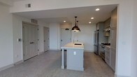 Located at 721 N High St in Columbus, Ohio, this single-floor space sits in the heart of the Short North Arts District, offering convenient access to the neighborhood's galleries, restaurants, and shops. The open-concept layout creates a sense of spaciousness, with the kitchen serving as a central focal point. A gray center island with a white countertop and an undermount sink anchors the kitchen, illuminated by a striking bronze pendant light that adds an industrial-modern touch. Recessed lighting throughout the ceiling provides consistent, even illumination across the space. The kitchen is equipped with stainless steel appliances, including a professional-grade gas range and a full-sized refrigerator, complemented by gray cabinetry that lines the perimeter wall. Speckled concrete or terrazzo-style flooring runs throughout, lending a durable and contemporary aesthetic to the space. White walls and white panel doors contribute to a clean, neutral palette, while multiple doorways suggest well-defined access points and functional flow. The overall ambiance is modern and minimalist, combining practical design with refined details suited to an urban setting.