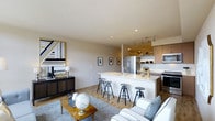 This thoughtfully designed single-floor space showcases a seamless open-concept layout that effortlessly connects the kitchen and living areas, creating an inviting and functional environment. The kitchen is a standout feature, equipped with stainless steel appliances, including a full-size refrigerator, microwave, and range. Warm wood-toned cabinetry lines the walls, complemented by track lighting that illuminates the space with a modern touch. A sleek peninsula with a light-colored countertop extends outward, offering seating for several bar-height stools — ideal for casual dining or entertaining. The living area flows naturally from the kitchen, anchored by a plush sectional sofa and a warm wood coffee table that adds character to the space. Light hardwood-style flooring runs throughout, enhancing the bright and airy atmosphere created by recessed ceiling lighting. Tasteful décor, including a bold geometric wall piece and framed artwork, adds personality and visual interest. The neutral color palette of soft whites and warm beiges lends a clean, contemporary feel throughout, making this a versatile and welcoming space suited for both everyday living and social gatherings.