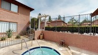 This outdoor spa area is part of a residential community characterized by warm terracotta-toned stucco walls and a classic tile-roofed architectural style. The centerpiece of the space is a circular hot tub finished with blue tile, equipped with stainless steel entry handrails and visible jet fixtures, offering a relaxing hydrotherapy experience. The spa is surrounded by a concrete deck with room for seating, including a small side table positioned nearby for convenience. An elegant white pergola structure serves as a decorative gateway entry point to the spa area, framed by mature trees and tall hedgerows that provide natural screening and privacy. The space is enclosed by a black iron perimeter fence, which opens to views of the surrounding community buildings and palm trees, lending a distinctly Southern California ambiance. Informational and safety signage is posted prominently on the stucco wall alongside a fire extinguisher station, noting spa rules, hours, and an occupancy limit, indicating the space is thoughtfully managed for resident safety. Mature landscaping, including leafy shrubs and ground-level plantings, adds greenery throughout the area. The overall setting is open-air and sun-drenched, making it a practical and pleasant communal amenity for residents to enjoy year-round.