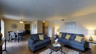 This single-floor residence in Denton offers a comfortable, open layout designed for everyday living. The living room serves as the central hub of the space, featuring hardwood-style flooring, a textured ceiling, and a ceiling fan that contributes to a casual, functional atmosphere. Two navy blue sofas with yellow accent pillows anchor the seating area around a dark coffee table, creating a cohesive and inviting gathering space. Decorative wall art adds a subtle touch of character to the neutral-toned walls.

The open floor plan allows a natural transition from the living area into the dining space, which is visible to one side and accommodates a dining table with seating. Beyond the dining area, the kitchen is accessible through a connecting corridor, featuring dark cabinetry that contrasts with the lighter surroundings. Ceiling-mounted light fixtures provide consistent illumination throughout the shared spaces. Interior doorways and hallways connect the various areas of the home seamlessly, maintaining an efficient and accessible flow. Overall, the space presents a clean, straightforward layout suited for comfortable day-to-day living.