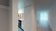 This single-floor unit offers a clean, modern layout designed for comfortable living. The spacious living area is finished with light-toned hardwood flooring that runs throughout, creating a warm and cohesive aesthetic. Large windows dressed with vertical blinds flood the space with natural light, contributing to an airy, open ambiance. A sliding glass door provides access to the outdoors, further enhancing the sense of openness.

The kitchen is efficiently designed with white cabinetry extending to the ceiling for ample storage, white countertops, and dark tile flooring that contrasts cleanly with the bright cabinetry. It is equipped with stainless steel appliances, including a refrigerator and microwave, as well as a black dishwasher. A pass-through window connects the kitchen directly to the living area, promoting an open flow between the two spaces.

The bedroom is generously proportioned, featuring the same light hardwood flooring as the living area, a large closet with paneled double doors, and floor-to-ceiling windows that bring in abundant natural light. A walk-in closet is also accessible from the bedroom, offering practical storage. Throughout the unit, white walls and clean finishes maintain a bright, neutral palette that suits a variety of interior styles.