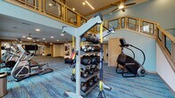 Located in Trophy Club, Texas, this two-story fitness center is a well-equipped facility designed to support a wide range of workout routines. The space makes a strong architectural impression with soaring double-height ceilings in the main area, floor-to-ceiling windows that flood the interior with natural light, and an open mezzanine level with warm wood-toned railings that overlooks the ground floor below.

The main level is expansive and thoughtfully organized into distinct workout zones. A dedicated cardio row lines one side of the space, outfitted with a generous selection of treadmills, ellipticals, and stair climbers. Nearby, a functional training area houses kettlebell racks, TRX suspension systems, battle ropes, and a StairMaster. Spin bikes are positioned beneath the tall windows, taking advantage of the abundant natural light and outdoor views.

A separate strength training zone features multiple cable machines, weight machines, squat racks, and free weight benches, complemented by full-length mirrors and ceiling fans throughout. The upper mezzanine level provides additional workout space and overlooks the main floor through open railings, creating a connected, airy feel. Throughout both levels, vibrant blue multi-tone carpeting adds a contemporary energy to the overall design.