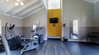 This fitness center offers a motivating and functional workout environment designed to support an active lifestyle. The space is defined by dramatic vaulted ceilings with exposed white beams that create an open, airy feel, allowing ample natural light to pour in through high clerestory windows. A bold yellow accent wall serves as a striking focal point, adding energy and vibrancy to the room while housing a wall-mounted flat-screen television for entertainment during workouts. The flooring transitions between durable rubber gym matting in the equipment zones and wood-style vinyl planking in the open stretching and exercise areas, providing both functionality and aesthetic appeal. Full-length mirrors are strategically positioned along the walls to enhance the sense of space and allow for proper form checks during exercise. The facility includes cardio equipment such as a stationary bike and an elliptical cross-trainer. Neutral grey tile wainscoting lines the lower portion of the walls, complementing the modern, clean design. Natural light from surrounding windows ensures the space remains bright and welcoming throughout the day, creating an inviting atmosphere for residents to pursue their fitness goals.