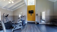 This fitness center offers a motivating and functional workout environment designed to support an active lifestyle. The space is defined by dramatic vaulted ceilings with exposed white beams that create an open, airy feel, allowing ample natural light to pour in through high clerestory windows. A bold yellow accent wall serves as a striking focal point, adding energy and vibrancy to the room while housing a wall-mounted flat-screen television for entertainment during workouts. The flooring transitions between durable rubber gym matting in the equipment zones and wood-style vinyl planking in the open stretching and exercise areas, providing both functionality and aesthetic appeal. Full-length mirrors are strategically positioned along the walls to enhance the sense of space and allow for proper form checks during exercise. The facility includes cardio equipment such as a stationary bike and an elliptical cross-trainer. Neutral grey tile wainscoting lines the lower portion of the walls, complementing the modern, clean design. Natural light from surrounding windows ensures the space remains bright and welcoming throughout the day, creating an inviting atmosphere for residents to pursue their fitness goals.
