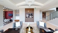 Located in Dallas, this single-floor space showcases a refined blend of contemporary design and upscale finishes. The kitchen area is a standout feature, boasting an expansive, high-gloss countertop that reflects the elegant pendant lighting above. Rich walnut cabinetry with open shelving frames a mosaic tile backsplash, complemented by stainless steel appliances, creating a sleek and functional culinary hub. A lounge area features a bold, patterned crimson accent wall with a wall-mounted flat-screen television, paired with floor-to-ceiling windows that frame sweeping city views. Ornate drum pendant chandeliers add warmth and drama to the space. A secondary sitting area draws attention with its striking coffered ceiling, featuring a geometric, multi-tiered design in crisp white. A wood-paneled accent wall serves as the focal point, housing another mounted television, while two sophisticated gray sofas and a metallic coffee table complete the refined look. A dining nook is defined by an upholstered white banquette with large-format padded wall panels, creating a cozy yet elegant atmosphere. Throughout, the space exudes a modern urban sensibility with a careful balance of bold accents and neutral tones.