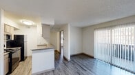 Located at 9900 Adleta Blvd in Dallas, TX, this one-bedroom, one-bathroom unit offers a practical and well-appointed single-floor layout. Hardwood-style flooring runs throughout the living areas, complementing the neutral wall tones and textured ceilings. The open-concept kitchen and living room area provides an efficient use of space, with the kitchen featuring a peninsula with a built-in sink, gray laminate countertops, light wood cabinetry, and black appliances including a range and refrigerator, alongside a stainless steel microwave. The living room receives ample natural light through a sliding glass door fitted with vertical blinds, which opens directly onto a private patio. The bedroom is generously sized and features multiple windows dressed with horizontal blinds, a ceiling fan with a light fixture, and hardwood-style flooring. Three closets throughout the unit offer notable storage capacity. The hallway connects the main living spaces to the bedroom and bathroom efficiently. The full bathroom includes a single-sink vanity with wood-toned cabinetry, a large mirror with overhead lighting, a toilet, and a tub-shower combination with tile surround. The entry area provides a clean transition point into the living space.