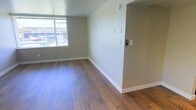 This single-floor space presents a clean, well-maintained interior designed for comfortable everyday living. The main living area is highlighted by warm, medium-toned hardwood-style flooring that spans the room, adding a sense of warmth and continuity throughout. Neutral, light beige walls provide a versatile backdrop that complements a variety of furnishing styles and décor choices. A large window along one wall allows generous natural light to fill the space, creating a bright and welcoming atmosphere. White baseboards and trim offer a crisp contrast against the flooring and walls, contributing to a tidy, finished look. Multiple light switches are conveniently grouped on the interior wall, and a security or intercom device is mounted nearby, adding a layer of functionality and safety to the unit. The textured ceiling adds subtle character overhead. The overall layout feels open and uncluttered, providing flexibility in how the space can be arranged and utilized. With its combination of natural light, durable flooring, and neutral finishes, this space offers a practical and inviting environment that is move-in ready.