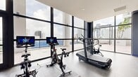 Located at 1642 NE 1st Ave in Miami, FL, this contemporary fitness center offers a well-appointed, expansive workout facility designed to meet a wide range of fitness needs. The gym's most striking architectural feature is its extensive floor-to-ceiling glass window walls, which flood the space with abundant natural light and frame sweeping views of the Miami city skyline and surrounding greenery, creating an energizing and motivating atmosphere.

The facility is organized into distinct functional zones across its generously sized single-floor layout. A dedicated cardio area houses multiple Stroops curved treadmills and stationary cycling bikes equipped with interactive display screens. Adjacent to this, a strength training section provides a comprehensive selection of selectorized machines, free weights, kettlebells, medicine balls, a punching bag, and a power rack with a bench press station. A turf sprint runner track with Aktiv branding adds a dynamic functional training element to the space.

The interiors feature a neutral palette of dark charcoal and muted blue accent walls, polished concrete-style flooring, and recessed ceiling lighting, all contributing to a clean, modern aesthetic. A glass-walled entrance corridor and a mirrored strength area enhance the sense of openness. Direct access to an outdoor terrace further distinguishes this gym as a premium fitness destination in the heart of Miami.