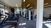 This modern fitness center is designed to provide a comprehensive workout environment within a single, thoughtfully appointed floor. The space is immediately distinguished by its eye-catching slatted wood ceiling, which adds warmth and architectural character to what might otherwise be a purely utilitarian setting. Recessed linear lighting runs along the ceiling, ensuring the entire area is brightly and evenly illuminated.

The fitness floor is generously laid out with a wide variety of equipment to support diverse workout routines. Along one side, a row of cardio machines — including treadmills and other aerobic equipment — lines the wall, taking advantage of large windows that invite natural light and provide views to the outdoors. Strength training is well represented, with a multi-station cable machine, a dedicated free weights section featuring medicine balls and dumbbells, and additional resistance equipment arranged for ease of use.

Large mirrors along the walls enhance the sense of space and allow users to monitor their form during exercise. The flooring consists of durable, low-pile carpet suited for a fitness environment, while the clean white walls and navy accent tones create a crisp, professional atmosphere. The overall ambiance is modern and motivating, balancing functional design with refined finishes.