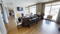 This single-story space showcases a thoughtfully designed living area that blends classic and contemporary elements. The room is anchored by rich hardwood flooring that extends throughout, providing a warm and inviting foundation. A prominent white-paneled fireplace serves as the focal point of the main seating area, flanked by a wall-mounted flat-screen television above. Large windows and glass-paned doors line the perimeter, flooding the interior with natural light while offering views of the surrounding exterior. Neutral-toned walls in warm beige and white complement the space's earthy color palette. The furnishings include a substantial dark leather sofa arrangement paired with rustic wooden console and side tables, accented by ceramic table lamps. Elegant floor-to-ceiling curtains in soft gray frame the windows, adding a refined layer of texture. Recessed lighting in the ceiling supplements the natural light, ensuring the space remains well-lit at all times. The overall ambiance is sophisticated yet comfortable, characterized by a harmonious mix of traditional architectural details and contemporary furnishings that create an welcoming, functional environment.
