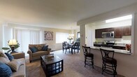 This single-floor residence offers an open-concept floor plan that seamlessly connects the living, dining, and kitchen areas into one cohesive and functional space. The living area features plush carpeting and is filled with natural light streaming through large windows dressed with vertical blinds, creating a bright and welcoming atmosphere. A comfortable seating arrangement anchors the space, complemented by a classic coffee table with built-in shelving.

The dining area sits centrally within the open layout, illuminated by an elegant chandelier that adds a refined touch to the space. It flows naturally from the living room, making it ideal for entertaining and everyday living.

The kitchen is designed with dark espresso-toned cabinetry, granite-style countertops, and stainless steel appliances, offering both style and practicality. A breakfast bar with bar stool seating provides a casual dining option and serves as a natural divider between the kitchen and the main living area.

Throughout the space, warm neutral wall tones and consistent flooring choices create a harmonious and inviting ambiance. The thoughtful open layout maximizes the sense of space, making this residence feel spacious, airy, and move-in ready.