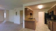 This single-floor space is designed with an open layout that connects the kitchen and living area seamlessly. The kitchen serves as a central feature, anchored by a peninsula with granite countertops and an integrated sink, offering both prep space and a casual seating option. Rich, dark wood cabinetry is mounted above, providing ample storage, while a neutral-toned tile backsplash adds texture and visual interest to the cooking area. The tile flooring in the kitchen transitions into carpeting in the adjacent living space, creating a clear distinction between the two zones while maintaining an open, connected feel. A flush-mounted ceiling light provides functional illumination over the kitchen area. The living space is open and unobstructed, with white walls and trim that lend a clean, neutral backdrop. A wall-mounted vent is visible, indicating a central HVAC system. Doorways visible from the main area suggest additional rooms beyond, offering practical separation of spaces. Overall, the space presents a straightforward, functional layout with classic finishes throughout.