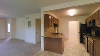 This single-floor space is designed with an open layout that connects the kitchen and living area seamlessly. The kitchen serves as a central feature, anchored by a peninsula with granite countertops and an integrated sink, offering both prep space and a casual seating option. Rich, dark wood cabinetry is mounted above, providing ample storage, while a neutral-toned tile backsplash adds texture and visual interest to the cooking area. The tile flooring in the kitchen transitions into carpeting in the adjacent living space, creating a clear distinction between the two zones while maintaining an open, connected feel. A flush-mounted ceiling light provides functional illumination over the kitchen area. The living space is open and unobstructed, with white walls and trim that lend a clean, neutral backdrop. A wall-mounted vent is visible, indicating a central HVAC system. Doorways visible from the main area suggest additional rooms beyond, offering practical separation of spaces. Overall, the space presents a straightforward, functional layout with classic finishes throughout.