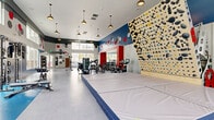 This expansive fitness and recreation facility is designed to support a wide range of athletic activities across several distinct zones. The centerpiece is a full-sized indoor basketball court with a polished hardwood floor, regulation hoops, and bold court markings, including a branded logo at center court. Tall windows line the court's perimeter, flooding the space with natural light and offering views of the surrounding outdoor areas. Padded wall panels provide safety along the court's sidelines, while a striking geometric mountain mural adds visual interest above.

Adjacent to the court, the fitness area features an impressive floor-to-ceiling bouldering wall densely set with colorful climbing holds, complemented by large crash mats positioned below for safety. Pendant lighting illuminates this zone, which also houses a well-equipped selection of strength machines, cable systems, free weights, and cardio equipment including stationary bikes and ellipticals. Open floor space with rubberized flooring accommodates functional training and stretching.

Clerestory windows and ceiling fans keep the fitness area bright and well-ventilated. Decorative hexagonal acoustic panels in red, blue, and gray add a dynamic, energetic aesthetic throughout. Entry is via a tiled vestibule with keycard access, ensuring a secure and polished arrival experience.