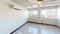 Studio B is a single-floor studio unit that combines an industrial aesthetic with functional living elements. The main living area is defined by polished concrete floors that extend throughout the space, exposed ceiling beams, and visible ductwork that contribute to the unit's loft-inspired character. A ceiling fan is centrally positioned, and a wall-mounted mini-split HVAC unit ensures year-round climate control. Large windows along one wall flood the space with natural light while framing expansive views of the surrounding urban cityscape, including notable city skyline and street-level activity below.

The kitchenette is efficiently designed, featuring white cabinetry, a stainless steel full-size refrigerator, an over-the-range microwave, and a single-basin sink with ample counter space. A dedicated closet area with sliding charcoal-toned doors provides generous storage, complemented by built-in shelving above. Industrial-style ceiling-mounted light fixtures illuminate the hallway and entryway.

The bathroom is cleanly appointed with a full tub and shower combination lined with white tile, a toilet, and a vanity. Rich, multi-toned tile flooring adds visual interest and contrasts with the crisp white walls, giving the bathroom a polished, practical finish.