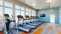 This single-floor fitness center is designed to support a comprehensive workout experience in a bright, open environment. The space is defined by large, multi-pane windows lining the walls, flooding the interior with natural light and offering views of the surrounding outdoor area. The color palette features calming blue accent walls paired with crisp white trim and ceilings, creating an energizing yet clean aesthetic. A ceiling fan is centrally mounted, complementing the flush-panel LED lighting above.

The flooring consists of modular carpet tiles in a gray wood-grain pattern accented by bold orange strips, adding visual energy and helping to delineate workout zones. A full-wall mirror on one side amplifies the sense of space and allows users to monitor their form during exercise.

The cardio zone is generously outfitted with a long row of Life Fitness treadmills positioned along the windowed wall, alongside stair climbers and elliptical machines. A wall-mounted flat-screen television provides entertainment during cardio sessions. The strength training area includes a multi-tiered dumbbell rack with benches, cable machines, and additional resistance equipment. The space also features a dedicated door providing access to an adjacent room or exit, ensuring smooth circulation throughout the facility.