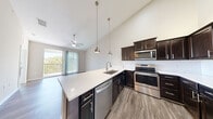 This well-appointed single-floor residence combines modern design with practical living across a thoughtfully laid-out floor plan. The kitchen serves as a central highlight, showcasing rich dark espresso cabinetry, white quartz countertops, a classic white subway tile backsplash, and a full suite of stainless steel appliances including a range, microwave, dishwasher, and refrigerator. Pendant lighting hangs from the dramatically vaulted ceilings, adding warmth and visual interest to the space. The kitchen flows seamlessly into the open living and dining area, where the soaring ceilings continue to create an airy, expansive atmosphere. Wide-plank wood-look flooring runs throughout the main living spaces, lending a cohesive and contemporary feel. A sliding glass door opens directly onto a private balcony with outdoor views, allowing natural light to pour into the living area. The primary bathroom features a corner vanity with matching dark cabinetry, white quartz countertops, a large framed mirror, and tile flooring. A dedicated in-unit laundry room provides added convenience, complete with a full-size washer and dryer and overhead wire shelving for storage. Neutral tones throughout the space create a clean, welcoming ambiance ideal for modern living.