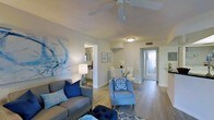 This single-floor residence offers a welcoming and thoughtfully designed living environment anchored by a coastal-inspired color palette of soft blues, whites, and warm neutrals. The open-concept layout seamlessly connects the living and dining areas, creating a sense of spaciousness and easy flow throughout the home. Light wood-tone flooring runs throughout the main living spaces, lending warmth and continuity to the design.

The living area is highlighted by a ceiling fan that enhances comfort, textured ceilings, and ample ambient lighting. A large, expressive blue abstract artwork serves as a striking focal point, complementing the curated furnishings including a plush grey sofa and a coordinating accent chair with blue medallion upholstery.

The dining area, positioned adjacent to the living room, features a glass-top table surrounded by white chairs, adding an elegant yet airy touch. A shelf niche in the dividing wall provides a subtle architectural detail that adds character to the space.

Natural light filters through sheer curtains visible toward the rear of the home, enhancing the bright and airy ambiance. A glimpse into what appears to be a well-lit bathroom with a vanity mirror further underscores the attention to detail carried throughout this thoughtfully finished residence.