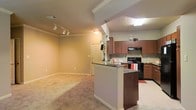 This single-story space showcases an open-concept layout that seamlessly connects the kitchen and living area. The kitchen is a standout feature, equipped with rich medium-toned wood cabinetry, dark granite countertops, and black appliances including a range and built-in microwave. A peninsula with matching granite overhang provides additional prep space and serves as a natural dividing point between the kitchen and adjoining areas. Pendant lighting hangs above the peninsula, complementing the tile flooring throughout the kitchen zone. The living area transitions to plush wall-to-wall carpeting, creating a warm and comfortable atmosphere. Track lighting installed along the ceiling offers flexible illumination across the open space. Crown molding adds a refined architectural detail along the upper walls, enhancing the overall finish of the interior. The layout is practical and well-proportioned, allowing natural sightlines throughout the space. Neutral wall tones throughout contribute to a clean, cohesive aesthetic that suits a variety of furnishing styles. An entry door is visible in the background, reinforcing the functional flow of the floor plan.