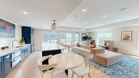 Located on the 4th floor of 601 Industrial Blvd W in Waconia, Minnesota, this thoughtfully designed clubroom serves as a premium shared amenity space. The open-plan layout flows seamlessly across light hardwood flooring, connecting distinct functional zones within a bright, airy environment. Recessed lighting and tray ceilings lend architectural interest overhead, while an abundance of large windows on multiple walls floods the space with natural light and frames views of the surrounding treetops and skyline.

The kitchenette area is a standout feature, showcasing bold navy cabinetry, white quartz countertops, a stainless steel sink, built-in refrigerator drawers, and a full-size stainless refrigerator. A striking scalloped tile backsplash serves as a decorative focal point beneath a large wall-mounted television, making it ideal for entertaining. A navy island with bar seating provides additional gathering space.

The lounge zone is anchored by a generous sectional sofa and accent chairs arranged atop a vibrant blue patterned area rug, creating a relaxed yet refined social setting. A dedicated game nook features a pool table set against a scenic landscape mural, adding a recreational dimension. Glass doors open directly onto an outdoor terrace equipped with patio furniture and string lights, extending the entertaining possibilities outdoors.