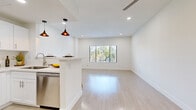 This single-floor space is designed with a clean, modern aesthetic defined by crisp white walls, light wood-tone flooring, and recessed lighting throughout. The open-concept layout connects the kitchen directly to the living area, creating a spacious, cohesive feel. The kitchen is well-appointed with white shaker cabinetry, stone countertops, stainless steel appliances including a refrigerator, range, microwave, and dishwasher, and a peninsula that doubles as a breakfast bar with seating. Warm amber pendant lights hang above the peninsula, adding a subtle contrast to the otherwise bright, neutral palette.

The living area is generously sized, featuring a ceiling fan and a window that frames views of surrounding greenery, drawing in ample natural light. Multiple bedrooms are present, each with large windows offering tree-lined outdoor views, light flooring, and recessed lighting. One bedroom features tall ceilings and a mirrored sliding-door closet, with direct access to a bathroom equipped with white cabinetry, a vanity mirror, and a full soaking bathtub. Throughout the space, the minimalist design, consistent materials, and abundant natural light contribute to a bright, airy, and cohesive living environment.