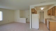 This single-floor space offers a straightforward, functional layout with a clean and neutral aesthetic throughout. The main living area is finished with wall-to-wall carpeting in a light, neutral tone, contributing to a warm and understated atmosphere. White walls and standard ceiling height give the space a simple, practical character.

A partial wall separates the living area from the kitchen, which is visible through an open passageway. The kitchen features wooden upper cabinetry in a medium honey tone, a full-size white refrigerator, and overhead lighting. An adjacent doorway suggests access to additional rooms, likely a bathroom or utility area, enhancing the functional flow of the space.

Toward the back of the living area, a staircase with a railing leads downward, indicating access to a lower level despite the single-floor classification, possibly a basement or storage level. This architectural detail adds depth to the layout.

Overall, the space presents a clean, move-in-ready environment with a neutral palette well-suited for a variety of furnishing styles. The open sightlines between the living and kitchen areas promote a connected, cohesive feel throughout the unit.