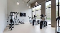 This fitness center offers a well-appointed, single-floor workout space designed with both functionality and comfort in mind. The room features notably high ceilings with an exposed concrete finish overhead, lending an industrial-modern aesthetic that feels open and energizing. Pendant-style industrial lights hang from above, providing warm, focused illumination throughout the space.

Large floor-to-ceiling windows line one wall, flooding the interior with abundant natural light and offering views of the surrounding outdoor greenery. The light-toned flooring complements the neutral gray walls, creating a clean and motivating environment.

The fitness center is equipped with a variety of cardio machines, including ellipticals and stationary bikes, providing options for a range of aerobic workouts. A multi-use strength and resistance training station is positioned along the main wall, alongside a wall-mounted flat-screen television — ideal for entertainment during workouts. The equipment is arranged to allow ample space for movement between stations. Overall, the space balances a minimalist, contemporary design with practical amenities suited for daily fitness use.