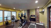 This single-level fitness room offers a well-equipped exercise environment distinguished by a standout exposed brick fireplace that serves as a dramatic architectural focal point along one wall. The warm yellow-green walls contrast with the rustic brick detailing, creating an inviting atmosphere uncommon in typical workout spaces. Recessed ceiling lighting provides even illumination throughout the room. The space is outfitted with cardio equipment including an elliptical trainer and treadmill, while a dedicated free weights area along one wall features a rack of dumbbells and a stability ball. A large mirror spans one wall, enhancing the sense of space and allowing users to monitor their form during exercise. A glass-paned door provides access to an adjacent outdoor area, introducing natural light into the room. Dark rubber flooring covers the exercise area, offering both durability and safety underfoot. The combination of functional fitness equipment, the unique brick fireplace, and thoughtful design details make this a distinctive and practical workout space.