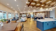 Located on Fisher Street in Charlottesville, Virginia, this single-floor community clubhouse offers a well-appointed gathering space defined by a blend of rustic and contemporary design elements. Warm hardwood flooring flows throughout, while exposed wood beam ceiling installations add architectural character to the expansive main lounge. Recessed lighting and pendant fixtures illuminate the open-plan layout, which features generous seating arrangements including a curved sectional sofa and patterned accent chairs, creating distinct conversation areas within a cohesive, airy environment.

Large windows along the perimeter admit abundant natural light and frame outdoor views of the surrounding Charlottesville landscape, with access to an exterior balcony visible from multiple areas. A boldly designed service counter with blue hexagonal tile detailing and white quartz countertops anchors one side of the main gathering space.

The kitchenette area includes matte gray cabinetry, white quartz counters, subway tile backsplash, stainless steel appliances, and a matte black fixtures. A private dining room accommodates a large farmhouse-style table beneath soaring ceilings. A dedicated business center provides individual workstations alongside lounge seating, while the game room features exposed beam ceilings, a foosball table, wall-mounted television, and direct balcony access.