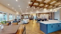 Located on Fisher Street in Charlottesville, Virginia, this single-floor community clubhouse offers a well-appointed gathering space defined by a blend of rustic and contemporary design elements. Warm hardwood flooring flows throughout, while exposed wood beam ceiling installations add architectural character to the expansive main lounge. Recessed lighting and pendant fixtures illuminate the open-plan layout, which features generous seating arrangements including a curved sectional sofa and patterned accent chairs, creating distinct conversation areas within a cohesive, airy environment.

Large windows along the perimeter admit abundant natural light and frame outdoor views of the surrounding Charlottesville landscape, with access to an exterior balcony visible from multiple areas. A boldly designed service counter with blue hexagonal tile detailing and white quartz countertops anchors one side of the main gathering space.

The kitchenette area includes matte gray cabinetry, white quartz counters, subway tile backsplash, stainless steel appliances, and a matte black fixtures. A private dining room accommodates a large farmhouse-style table beneath soaring ceilings. A dedicated business center provides individual workstations alongside lounge seating, while the game room features exposed beam ceilings, a foosball table, wall-mounted television, and direct balcony access.