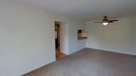 This single-floor space presents a straightforward and functional layout centered around an open living area. The room is finished with neutral-toned carpet flooring and white walls, creating a clean and bright atmosphere. A ceiling fan with a light fixture is mounted overhead, providing both air circulation and ambient lighting throughout the space.

A key architectural feature is the pass-through counter that connects the living area to the kitchen. This built-in counter, finished in warm wood tones, offers a convenient serving or casual dining surface while maintaining an open sightline between the two spaces. The kitchen, visible through both the pass-through and an open doorway, features hardwood or laminate flooring and wood-toned cabinetry, offering a visual contrast to the carpeted living area.

The open doorway leading into the kitchen enhances the flow and connectivity between the rooms, making the overall layout feel accessible and practical. White baseboards frame the carpeted floors throughout. The overall ambiance is neutral and move-in ready, offering a functional living space with a simple, understated design aesthetic suited to a variety of interior styles.