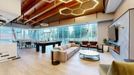 Located at 1915 Sooke Rd in Colwood, BC, this single-floor amenity lounge is a thoughtfully designed communal space that blends contemporary style with warm, natural materials. The room is anchored by striking exposed wood beam ceilings that rise to impressive heights, complemented by eye-catching honeycomb and circular pendant light fixtures that add a sculptural quality to the space overhead. Light wood-toned flooring runs throughout, contributing to an airy, welcoming atmosphere.

Floor-to-ceiling glazing spans a full wall, flooding the interior with abundant natural light and offering unobstructed views of the surrounding mature trees — creating a seamless connection between the indoors and the natural landscape outside. A set of glass doors leads to an outdoor patio area, extending the usable space further.

The lounge is organized into distinct functional zones. A comfortable seating area features a neutral sectional sofa and plush armchairs arranged around a sleek fireplace with a large-format TV mounted above a marble-clad surround. A games area includes a full-size pool table and foosball table. A generous kitchen bar with a waterfall-edge marble countertop, bar seating, and built-in cabinetry with dual screens serves as an entertainment hub. Dining tables with upholstered seating provide additional gathering space, completing this versatile and well-appointed amenity room.
