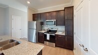 Located near I-35W N in Shoreview, Minnesota, this single-floor unit offers a thoughtfully designed open-concept layout with clean finishes throughout. The kitchen serves as a central highlight, featuring rich dark cabinetry, granite countertops, a large center island with a double-basin sink, and a full suite of stainless steel appliances including a refrigerator, range, and over-the-range microwave. Recessed lighting and crown molding add a polished touch to the space, while light hardwood-style flooring flows seamlessly through the main living areas. The open living area is generously proportioned, bathed in natural light and offering a bright, airy feel with neutral gray walls and crisp white trim. A dedicated bedroom features plush carpet, a wide window with outdoor views, and access to a spacious walk-in closet outfitted with multi-tier wire shelving systems for organized storage. The bathroom is well-appointed with a dark wood vanity, granite countertop, and a tub-shower combination, accented by a window that draws in natural light. A carpeted stairwell area with a handrail and additional window adds architectural interest. The unit combines functional design with modern finishes, making it a comfortable and stylish living space in Shoreview.