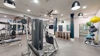 Located at 2800 Pontiac Dr in Aurora, Illinois, this well-appointed fitness center offers a generously sized, single-floor exercise space designed to support a comprehensive workout experience. The open floor plan is covered in durable speckled rubber flooring, providing both safety and comfort underfoot throughout the space.

The cardio zone is equipped with multiple treadmills, stair climbers, stationary bikes, and spin cycles, catering to a wide range of aerobic training preferences. Strength training is well represented with a multi-station cable and resistance machine, free weights, and a medicine ball rack stocked with various weighted balls for functional fitness exercises.

The interior design features a clean, modern aesthetic with white walls accented by bold navy blue columns and a striking accent wall. Pendant lighting and ceiling fans are suspended throughout, ensuring consistent air circulation and a well-lit environment. Full-length mirrors line a section of the wall, amplifying the sense of space and allowing users to monitor their form during workouts.

A wall-mounted flat-screen TV provides entertainment, while a built-in wood cubby storage unit offers convenient organization. A water fountain is also available for hydration. Large windows along one side of the room invite natural light, and high clerestory windows contribute to the bright, energetic atmosphere of this comprehensive fitness facility.