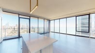 Located in Chicago's South Loop at 1200 S Indiana Ave, this high-rise unit offers a refined modern living experience defined by sweeping, unobstructed views of Lake Michigan and the Chicago skyline. Floor-to-ceiling glass walls wrap around the open living and dining areas, flooding the space with abundant natural light and framing breathtaking vistas of the waterfront and city below.

The living area features wide-plank wood flooring and a clean, open layout that maximizes the dramatic outlook. A gold-accented pendant fixture highlights the kitchen island, which is topped with light stone and pairs seamlessly with the custom cabinetry finished in a warm gray wood grain. The kitchen is equipped with Bosch stainless steel appliances, a gas cooktop, built-in oven, integrated refrigerator, and a white stone backsplash, combining function with understated elegance.

The bathrooms are thoughtfully finished with light wood vanities, dark stone countertops, large-format terracotta floor tiles, and glass-enclosed walk-in showers clad in textured tile. A sliding barn-style door adds a contemporary detail to the interior hallway, which connects the private rooms with clean white walls and recessed lighting throughout.