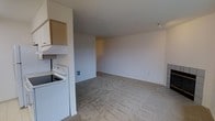 This single-floor space presents an open, functional layout that combines the kitchen and living areas into one continuous space. The kitchen is positioned along one wall and includes a white electric range, an overhead microwave or range hood, and light wood cabinetry mounted above for storage. The transition from the kitchen into the main living area is seamless, with the space opening up generously to accommodate a variety of furniture arrangements. Flooring throughout is wall-to-wall carpet in a neutral beige tone, lending a uniform, understated look across the space. The walls are painted a clean, light cream white, contributing to a bright and airy atmosphere. A built-in wall outlet and a track light fixture on the ceiling are among the practical details visible. The layout is straightforward and efficient, making good use of the available footprint. The overall ambiance is clean and minimal, offering a blank canvas well-suited for personalization. The single-level design ensures easy navigation throughout, making it a practical and accessible living space.