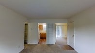 This single-floor residential unit offers a practical and functional layout designed to maximize living comfort. The main living area is finished with wall-to-wall carpeting, creating a warm and inviting atmosphere. From this central space, the layout opens to multiple areas, providing a sense of flow and connectivity throughout the unit.

The kitchen is accessible directly from the living area and features warm wood-toned cabinetry, a white appliance suite, and a window above the sink that allows natural light to fill the space. A built-in shelving unit between the living area and the adjoining room serves as both a functional storage solution and an open divider, adding architectural character to the interior.

The bedroom is bright and airy, benefiting from a large vertical window with vertical blinds that filter in generous amounts of natural light. Carpeted flooring continues into this room, maintaining a consistent and comfortable feel. A closet with sliding doors provides ample storage.

Additional built-in shelving near the entryway offers practical storage from the moment you step inside. The unit's neutral color palette throughout — white walls and light-toned finishes — creates a clean, open ambiance that is easy to personalize. Overall, the space is well-organized, efficiently designed, and filled with natural light.