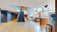 This expansive two-story communal facility offers a thoughtfully designed blend of industrial and contemporary architecture across two generously sized floors. The ground floor makes a striking first impression with its dramatic double-height atrium, polished concrete floors, and a bold stacked-stone fireplace anchoring the central lounge area. A sleek staircase with metal railings connects the two levels, while large windows flood the space with abundant natural light and outdoor views. The lounge is furnished with vibrant, colorful seating arrangements — including bold blue sofas, chartreuse accent chairs, and patterned ottomans — creating an energetic yet welcoming atmosphere. A reception or bar area with warm wood paneling adds a refined touch near the entry. The ground floor also features a game and social zone with high-top seating, arcade games, foosball, and a large-screen media area with a kitchenette. The upper floor showcases soaring exposed timber roof trusses and open truss ceilings that lend a warm, loft-like character. This level is organized into flexible lounge and collaborative seating zones, with hardwood flooring, abundant greenery, open shelving, and a variety of seating configurations suited for both casual gatherings and focused work. Throughout both floors, exposed ductwork, pendant lighting, and curated artwork contribute to a modern, creative ambiance.