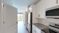 Located at 630 Washington Ave in Belleville, NJ, this thoughtfully designed studio offers a efficient, single-floor layout combining a kitchen and living area with one full bathroom. The kitchen is outfitted with white shaker-style cabinetry featuring matte black hardware, gray quartz countertops, a hexagonal tile backsplash, and a full suite of stainless steel appliances including a refrigerator, electric range, microwave, and dishwasher. A louvered interior door adds a distinctive architectural detail near the entry. The open living area is bathed in natural light through expansive floor-to-ceiling windows, which frame views of an outdoor pool and surrounding greenery. Light-toned hardwood flooring runs throughout, complementing the clean, neutral palette of the space. A wall-mounted mini-split HVAC unit provides efficient climate control. Built-in shelving, drawers, and a desk area offer practical storage and workspace solutions. The bathroom features large-format light tile flooring, a glass-enclosed walk-in shower with matte black fixtures, and a clean, minimalist finish. Recessed lighting throughout reinforces the bright, contemporary ambiance of the entire space.