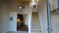 This two-story loft-style space is defined by its dramatic vertical volume, with soaring double-height ceilings that create an airy, open atmosphere on the main level. A ceiling fan is mounted high above, serving both the lower living area and enhancing airflow throughout the space. The main floor features dark hardwood-style flooring that anchors the open-concept layout, where the kitchen sits prominently along one wall. The kitchen is equipped with stainless steel appliances, including a refrigerator, stove, and dishwasher, complemented by dark cabinetry with modern hardware and a sleek countertop with an integrated sink. Track lighting illuminates the kitchen area efficiently. A carpeted staircase with a clean white railing leads to the upper loft level, which overlooks the main floor below. The upper level features a built-in closet with shelving and rod storage, making it a functional secondary space. The two-tone wall color—neutral gray tones with bright white trim—gives the interior a contemporary, cohesive feel. The overall design balances industrial-loft character with practical residential amenities.