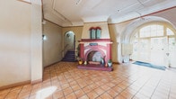 This single-floor lobby space makes a strong first impression through its distinctive architectural character and Mediterranean-inspired design elements. The centerpiece of the room is a striking decorative fireplace clad in red brick with arched detailing and a tiled surround, flanked by potted plants that add a welcoming touch. Warm terracotta tile flooring extends throughout the entire space, contributing to the overall cohesive aesthetic.

Arched doorways and vaulted ceiling sections are prominent throughout, lending the lobby a sense of elegance and Old World charm. A large arched window and double French doors on one end flood the space with natural light and offer views of an outdoor courtyard, where mature trees and seating areas are visible. The entry is framed by a grand arched transom window above the main door, reinforcing the architectural theme.

Residents are served by two banks of individual mailboxes mounted along the walls near the entrance. A staircase accessible through an arched opening leads to additional areas of the building. The space also includes utility areas with mailbox infrastructure and access doors. Exposed ceiling pipes in one section hint at the building's utilitarian underpinning, balanced by the warmth of the overall design. The lobby presents a functional yet characterful communal space with ample natural light and distinctive architectural personality.