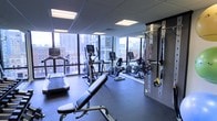 This fitness center offers a well-appointed exercise space designed to support a comprehensive workout experience. The room is anchored by large, floor-to-ceiling windows that flood the space with natural light while providing impressive views of the surrounding urban skyline, creating an energizing backdrop for any workout.

The cardio zone is equipped with multiple machines, including treadmills, ellipticals, and a recumbent bike, all positioned to take full advantage of the panoramic city views. A dedicated strength and functional training area lines one wall, featuring a wall-mounted cable and resistance system along with TRX suspension trainers, offering versatile options for full-body conditioning. An adjustable weight bench provides additional flexibility for a variety of strength exercises.

The space is finished with a dark rubberized flooring, ideal for both equipment stability and ease of maintenance. Recessed panel lighting complements the abundant natural light from the windows, ensuring the room remains bright and welcoming at any hour. The overall design strikes a balance between functionality and a clean, modern aesthetic, making this fitness center a well-equipped and visually engaging environment for exercise.