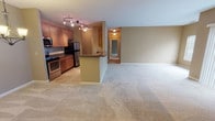 This single-floor residence offers a thoughtfully designed open-concept layout that seamlessly connects the kitchen and living areas. The kitchen is well-equipped with warm wood cabinetry, dark granite countertops, stainless steel appliances including a refrigerator, stove, and microwave, and track lighting overhead. A peninsula with a built-in sink serves as a natural divider between the kitchen and the expansive carpeted living and dining area, which is illuminated by a classic chandelier and features large vertical-blind-covered windows that allow generous natural light to fill the space. The warm neutral tones on the walls throughout the unit create a cohesive and inviting ambiance. The living area opens to a hallway leading to the private quarters, including generously sized bedrooms with plush carpeting and additional window access. The unit includes two bathrooms: one featuring a tiled walk-in shower with sliding glass doors and granite vanity, and the other offering a full soaking tub with decorative tile surround. Tile flooring in the kitchen and bathrooms provides a practical contrast to the soft carpeting found throughout the main living spaces and bedrooms.