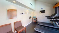 This fitness center is a thoughtfully designed, single-floor exercise space that provides a well-rounded workout environment. The room features a dark rubberized floor, white walls, and elevated ceilings with recessed lighting, contributing to an open and functional atmosphere. A wall-mounted flat-screen television keeps users entertained during cardio sessions.

The cardio area is anchored by a row of treadmills positioned along large windows and a glass door, offering direct views of a sparkling outdoor pool and surrounding courtyard — a motivating backdrop for any workout. Adjacent to the cardio zone, an elliptical trainer and stationary bike occupy a naturally lit corner, with windows overlooking a landscaped exterior and warm wall sconces providing ambient light.

A dedicated stretch and flexibility area houses stability balls and a wall-mounted mirror, ideal for warm-ups and cool-downs. Nearby, a shelving unit organizes kettlebells, resistance bands, yoga mats, and other small accessories for varied training routines. A wall-mounted shelf displays informational materials and motivational items, while a seating chair provides a rest area.

Facility rules are posted prominently, and a hand sanitizer dispenser and fire extinguisher reflect attention to safety and hygiene. The space benefits from abundant natural light streaming through multiple windows, creating a bright and inviting environment for residents to maintain an active lifestyle.