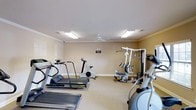 This fitness center offers a practical and well-organized space designed to support a well-rounded workout routine. The room features neutral beige walls accented by clean white crown molding and baseboards, lending a polished and inviting atmosphere. Overhead, flush-mounted fluorescent light panels provide bright, even illumination throughout the space, complemented by a ceiling fan that helps maintain air circulation during workouts. The flooring is carpeted, providing a comfortable surface underfoot. The equipment selection covers both cardiovascular and strength training needs. Multiple treadmills are positioned along one side of the room, alongside a stationary upright bike, offering a variety of cardio options. A multi-station cable and weight machine anchors the strength training area, providing users with options for a wide range of resistance exercises. An elliptical trainer rounds out the equipment, offering a low-impact cardio alternative. The layout allows for comfortable movement between stations, making the space functional for individual use. Overall, this single-floor fitness center is thoughtfully arranged to deliver an accessible and efficient exercise environment.