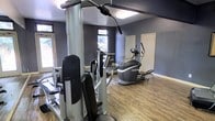 This fitness center offers a practical and functional workout environment designed to support a range of exercise routines. The space features wood-look flooring throughout, providing a clean and durable surface well-suited for gym use. The walls are painted in a deep blue-grey tone, giving the room a focused and energetic atmosphere. Natural light filters in through multiple windows along the walls, creating a bright and welcoming environment. The equipment selection includes a multi-function cable and weight machine for strength training, a treadmill for cardiovascular workouts, and an elliptical trainer for low-impact cardio exercise. The layout allows for comfortable movement between machines without feeling cramped. Overhead fluorescent lighting supplements the natural light, ensuring the space remains well-lit throughout the day. A white door provides access to an adjoining area. The overall design is straightforward and purpose-driven, prioritizing usability and functionality for residents or members looking for convenient access to fitness amenities.